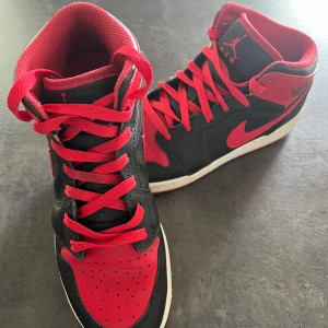 Nike Air Jordan 1 Svart/Röd Highs - Snygga Nike Air Jordan 1 i klassisk svart och röd färg med högt skaft. Skorna har röda snören, svart läder och röda detaljer på tå, sula och logga. Perfekta för dig som vill sticka ut med en ikonisk sneaker.