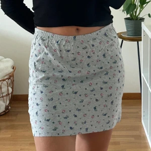 Grå blommig kortkjol med resår - Superfin grå kortkjol med små blommor i rosa och blått. Kjolen har elastisk midja för skön passform och är gjord i mjuk bomull. Perfekt för dig som gillar en söt och avslappnad stil.