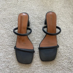Svarta sandaler med klack och fyrkantig tå - Stilrena svarta sandaler med fyrkantig tå och två tunna remmar över foten. Skorna har en låg blockklack och är tillverkade i ett material som liknar skinn. Perfekta för dig som gillar minimalistisk och trendig design.