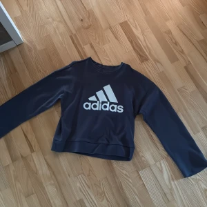Mörkblå Adidas sweatshirt - Snygg mörkblå sweatshirt från Adidas med stort vitt logotryck på bröstet. Tröjan har en klassisk passform, rund halsringning och långa ärmar. Perfekt för en sportig och avslappnad stil.