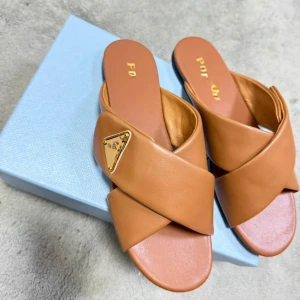 Bruna sandaler från Prda i skinn - Snygga bruna sandaler från Prda med korslagda remmar i mjukt skinn och guldfärgad logodetalj på sidan. Platt sula och öppen tå ger en clean och stilren vibe. Perfekta för varma dagar när du vill ha både komfort och lyxig känsla.