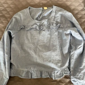 Ljusblå blus med volangdetaljer - Supersöt ljusblå blus med volanger både framtill och längs nederkanten. Blusen har rund halsringning, lång ärm och en liten knapp i nacken. 