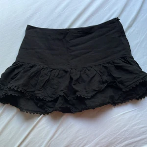 Svart volangkjol från Zara - Supersöt svart kortkjol från Zara med dubbla volanger och spetsdetaljer längst ner. Kjolen har dragkedja i sidan och är i ett mjukt bomullstyg. Perfekt för dig som gillar en feminin och trendig look.