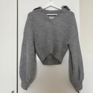 Grå croppad stickad hoodie Chiquelle - Supermysig grå stickad hoodie från Chiquelle i croppad modell. Tröjan har v-ringning, långa ärmar med ballongform och ribbade muddar. Perfekt för lager-på-lager och chill dagar. Snygg och enkel att styla till jeans eller kjol.