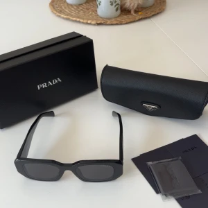 Svarta solglasögon från Prada - Snygga svarta solglasögon från Prada med breda skalmar och cool, kantig form. Glasögonen har mörka linser och tydlig Prada-logga på sidan. Kommer med originaletui och putsduk. Perfekt accessoar för en trendig look. 