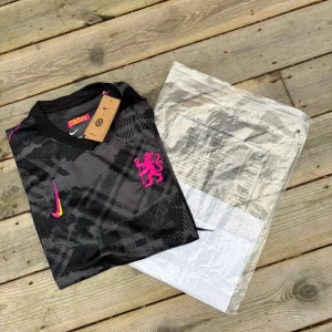Chelsea FC fotbollströja  - Grym svart fotbollströja från Nike med coolt grafiskt mönster och neonrosa detaljer. Tröjan har korta ärmar, V-ringning och en rosa lejonlogga på bröstet. Swoosh-loggan är i gult och rosa. Materialet är lätt och andas, perfekt för match eller träning. Pris kan diskutera vid snabb affär 