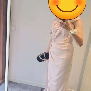 Ljusrosa långklänning med spets - Elegant ljusrosa långklänning med spetsdetaljer upptill och omlottkänsla i kjolen. Klänningen har offshoulder-skärning och vackra draperingar som ger en lyxig känsla. Perfekt för bal eller student.