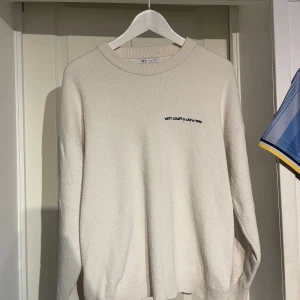 Beige stickad tröja från Zara - Säljer en beige stickad tröja från Zara med broderad text 'West County Crew 1990' på bröstet. Tröjan har rund halsringning och långa ärmar, perfekt för lager-på-lager och chill stil. Enkel och clean design som funkar till det mesta.