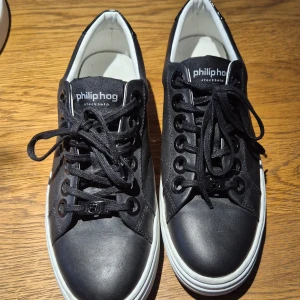 Svarta sneakers från Philip Hog, strl 39 - Stilrena svarta sneakers från Philip Hog med vita sulor och svarta snören. Skorna har rund tå, platt sula och är tillverkade i skinn. Snygga detaljer med logga på häl och snörning. Perfekta för dig som gillar en clean och modern look. Äkta skinnskor, använda 1 gång, ny skick. Ny pris 1100