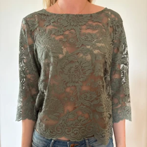 Grön spetsblus med trekvartsärm - Snygg grön blus i genombruten spets med stora blommönster och trekvartsärmar. Blusen har rund halsringning och vågig kant nertill och vid ärmslut. Perfekt för dig som vill ha en romantisk och trendig look.