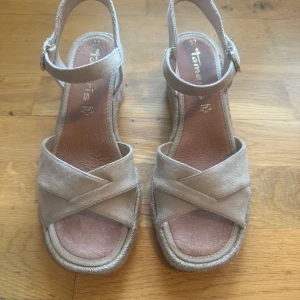 Beige mockasandaler med kilklack från Tamaris storlek 37 - Snygga sandaler från Tamaris i beige mocka med korsade remmar och justerbart spänne runt ankeln. Aldrig använda, köpta på eurosko för 800:-