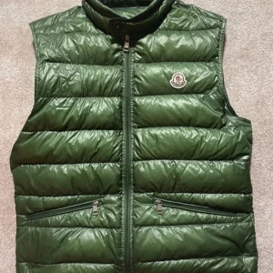 Grön dunväst från Moncler - Snygg grön dunväst från Moncler med dragkedja och hög krage. Västen har två praktiska fickor med dragkedja framtill och Monclers logga på bröstet. Perfekt för kyliga dagar.