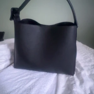 Svart axelväska i skinn - Snygg svart axelväska i skinn med stilren design. Väskan har en bred axelrem och en praktisk dragkedja på insidan för säker förvaring. Perfekt för att bära med sig det nödvändigaste med stil.