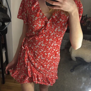 Röd blommig omlottklänning - Snygg röd omlottklänning med blommigt mönster och volangdetaljer. Klänningen har korta ärmar och en knytning i midjan som ger en fin passform. Perfekt för sommarens alla tillfällen💞