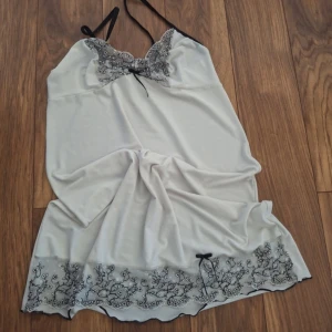 Nattlinne  - Elegant vit nattlinne med svarta spetsdetaljer vid halsringningen och nederkanten. Axelbandslös design med tunna svarta axelband och en liten rosett framtill. Perfekt för en lyxig känsla.