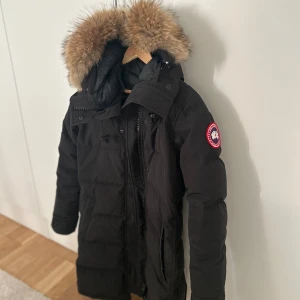 Svart dunjacka från Canada Goose - Säljer en svart dunjacka från Canada Goose med en stor, fluffig päls på huvan. Jackan har en dragkedja och knappar framtill samt en logotyp på ärmen. Perfekt för kalla vinterdagar.         Kvitto finns från NK!  Storlek S