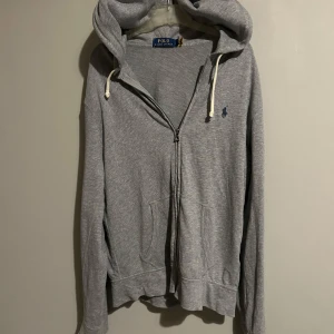 Grå hoodie från Polo Ralph Lauren - Säljer en stilren grå hoodie från Polo Ralph Lauren med dragkedja och broderad logga på bröstet. Tröjan har en justerbar huva med snören och praktiska fickor framtill. Perfekt för en avslappnad look. storlek s!