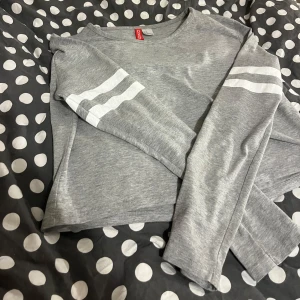 Grå croppad tröja  - Säljer en grå croppad tröja från Divided med vita ränder på ärmarna. Tröjan har långa ärmar och en avslappnad passform. Perfekt för en casual look!