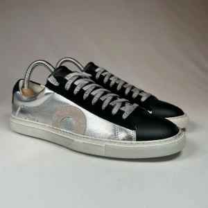 Svarta och silverfärgade sneakers från Oliver Cabell - Snygga sneakers i svart och silver från Oliver Cabell. Skorna har en unik design med ett stort cirkelmönster på sidan och märkesnamnet på snörena. Perfekta för dig som vill sticka ut med stil. 9,5/10 skick. 