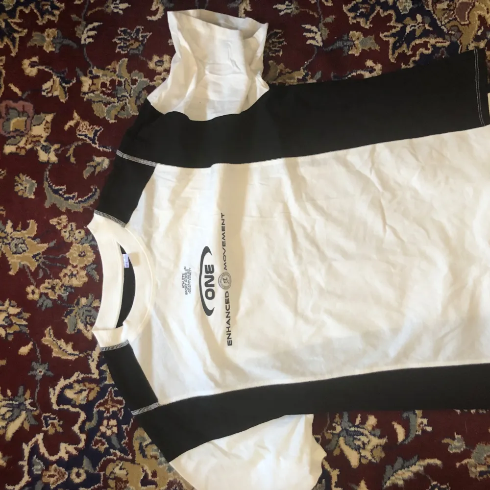 Säljer en vit och svart t-shirt med tryck 'ONE ENHANCED MOVEMENT' på framsidan. T-shirten har korta ärmar och en rund halsringning. Perfekt för en sportig look eller avslappnad stil.. T-paidat.
