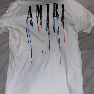 Vit t-shirt från Amiri med  - Säljer en vit t-shirt från Amiri med coola färgstänk och logotyp på både fram- och baksidan. Perfekt för en stilren men ändå unik look. T-shirten har korta ärmar och är gjord i mjukt material.