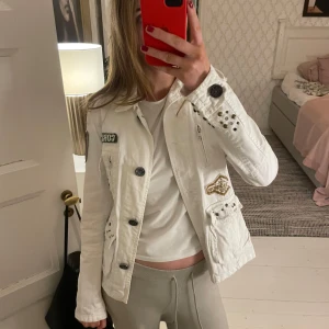 Vit jacka med nitar och patchar - Säljer en cool vit jacka med unika detaljer som nitar och patchar. Jackan har dragkedja och knappar, samt flera fickor. Perfekt för dig som vill sticka ut med en edgy stil. Storleken är inte angiven, men skulle säga att det är en S