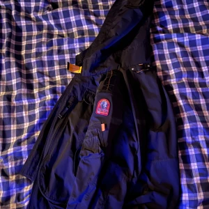 Svart jacka från Parajumpers - Säljer en svart jacka från Parajumpers med coola detaljer som en broderad logga på ärmen och meshfoder. Jackan har en dragkedja och flera praktiska fickor. Perfekt för dig som vill ha en stilren och funktionell jacka. Jackan är extremt fräsch överlag och har inga defekter. OBS! Juniormodel M