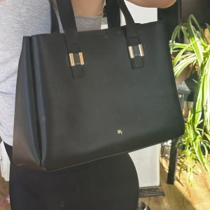 Svart axelväska med gulddetaljer - Snygg svart axelväska i skinn med eleganta gulddetaljer. Väskan har två rejäla handtag och en praktisk dragkedja inuti för säker förvaring. Perfekt för att bära med sig det nödvändigaste med stil.