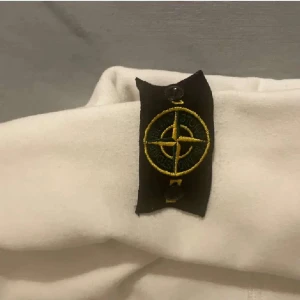 Vit hoodie med dragkedja från Stone Island - Säljer en stilren vit hoodie från Stone Island med dragkedja och huva. Den har en ikonisk logga på ärmen och praktiska fickor framtill. Perfekt för en avslappnad stil.