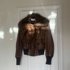 Brun skinnjacka med pälsdetalj från Marlboro Classics - Snygg brun skinnjacka från Marlboro Classics med en lyxig pälsdetalj runt kragen. Jackan har långa ärmar och ribbade muddar vid ärmslut och nederkant. Perfekt för att hålla stilen i kyligare väder!