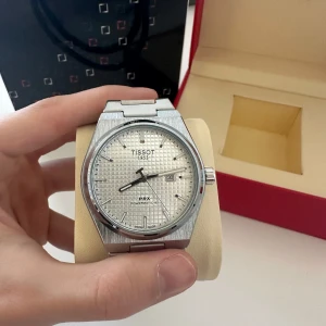 Tissot PRX Klocka - Snygg och stilren klocka från Tissot med silverfärgad boett och armband. Urtavlan har ett diskret rutmönster och visar datum. Perfekt för den som gillar klassisk design med en modern touch.