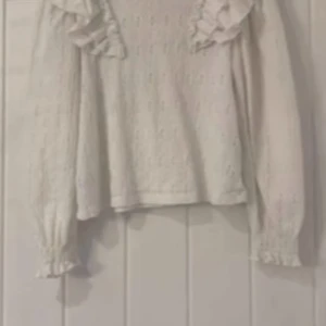 Vit stickad tröja med volangdetaljer - Säljer en söt vit stickad tröja med volangdetaljer vid axlarna. Tröjan har långa ärmar och ett diskret mönster som ger en fin textur. Perfekt för en stilren och mysig look.