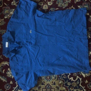 Blå pikétröja från Lacoste - Säljer en klassisk blå pikétröja från Lacoste med den ikoniska krokodilloggan broderad på bröstet. Tröjan har korta ärmar och en traditionell krage med knappar. Perfekt för en stilren och avslappnad look.