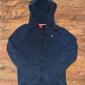 Mörkblå hoodie från Nike - Säljer en mörkblå hoodie från Nike med dragkedja och huva. Tröjan har en liten Nike-logga på bröstet och är perfekt för en avslappnad stil. Den har långa ärmar och är tillverkad i ett mjukt material.