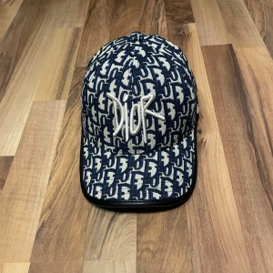 Mönstrad keps från Dior - Snygg keps från Dior med ett iögonfallande mönster i blått och beige. Kepsen har en broderad logotyp framtill och justerbar rem baktill för perfekt passform. Perfekt för att ge din outfit en lyxig touch.