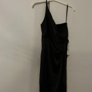 Svart one shoulder klänning från Bubbleroom - Snygg svart one shoulder klänning från Bubbleroom. Perfekt för en kväll ute med sin eleganta design och smickrande passform. Klänningen har en stilren look med en axelbandslös stil som ger en modern touch.