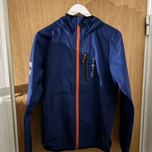 Blå vindjacka sail racing - Säljer en blå vindjacka från Gore-Tex med en iögonfallande orange dragkedja. Jackan har en justerbar huva och en praktisk bröstficka med dragkedja. Perfekt för utomhusaktiviteter och skyddar mot vind och regn.