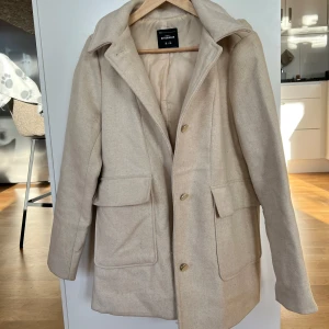 Beige vårjacka med luva - Säljer denna kappan köpt i Portugal. Stilren rak modell med gulknappar, perfekt till våren. Endast använd ett fåtal gånger