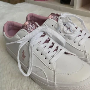 Vita sneakers från Polo Ralph Lauren - Säljer dessa supersnygga sneakers från ralph lauren!!💗💗 Skorna har aldrig använts och är därför i jättebra nyskick!!🩷 Storlek 36, men skulle säga att de är lite större i storlek och passar bättre som 37🩷🩷 Skorna är vita med rosa glittriga detaljer!✨