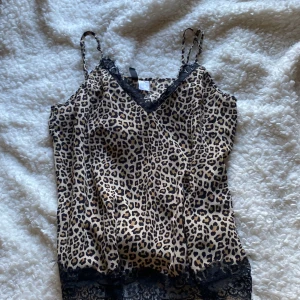 Leopardmönstrad linne med spetsdetaljer - Snygg leopardmönstrad linne med svarta spetsdetaljer vid halsringning och nederkant. Tunna axelband och v-ringad design ger en elegant touch. Perfekt för att lägga till lite vild stil i garderoben 🐆