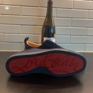 Mörkblå sneakers från Louboutin - Snygga mörkblå sneakers från Louboutin med den ikoniska röda sulan. Skorna har en stilren design med snörning och är tillverkade i mocka. Perfekta för dig som vill ha en touch av lyx i vardagen.