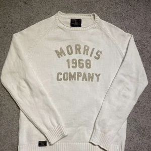 Vit stickad tröja från Morris - Säljer en stilren vit stickad tröja från Morris med texten 'Morris 1968 Company' i beige på framsidan. Tröjan har långa ärmar och ribbade muddar vid ärmslut och nederkant. Perfekt för en avslappnad och trendig look.