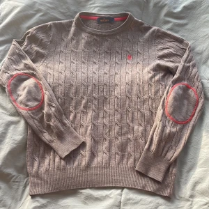 Beige Morris tröja - Säljer en snygg beige stickad tröja från Morris med kabelstickat mönster och röda detaljer på armbågarna. Tröjan har en broderad röd logga på bröstet och långa ärmar. Perfekt för kyligare dagar!