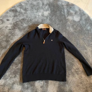 Mörkblå tröja med dragkedja från Morris - Snygg mörkblå tröja från Morris med dragkedja och en liten broderad detalj på bröstet. Tröjan har en kontrasterande krage i beige och långa ärmar. Perfekt för en stilren look.