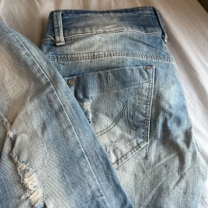 låga ljusa slitna bootcut jeans - Snygga ljusblå jeans med slitningar och en bootcut-stil. De har en låg midja och är perfekta för en avslappnad look. Jeansen har klassisk femficksdesign och knappar i midjan. Märket är LTB men byxorna är köpte secondhand.