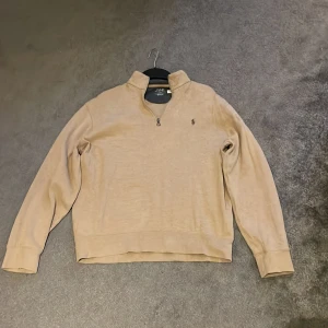 Beige tröja från Ralph Lauren - Säljer en stilren beige tröja från Ralph Lauren med dragkedja vid halsen. Tröjan har långa ärmar och en liten broderad logga på bröstet. Perfekt för en avslappnad och snygg look.