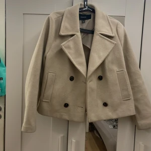 Beige kappa från VERO MODA - Säljer en stilren beige kappa från VERO MODA med dubbelknäppning och breda slag. Jackan har en klassisk design med två framfickor och är perfekt för höst och vår. Den är tillverkad i ett mjukt material som ger en elegant look. Kostar 699kr annars 