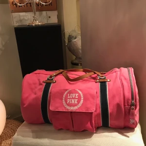 Rosa duffelväska från PINK - Säljer en snygg rosa duffelväska från PINK med texten 'LOVE PINK' på framsidan. Väskan har bruna handtag och en praktisk dragkedja. Insidan är mönstrad med ränder och logotyper. Perfekt för resor eller gymmet! Väskan är aldrig använd men axelremfästet var trasigt från butik. Går att du för den händige. De andra handtagen ( som syns) funkar utmärkt. 