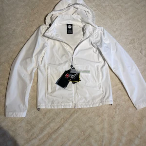 Vit jacka från Colmar (HELT NY) - Säljer en stilren vit vindjacka från Colmar. Jackan har en praktisk dragkedja framtill och en huva för extra skydd. Perfekt för blåsiga dagar och tillverkad av återvunnet material. Märkeslogga på ärmen ger en snygg touch. Helt ny, skick 10/10