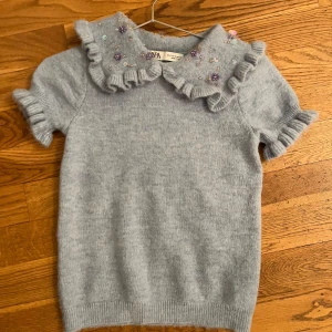 Ljusblå tröja med volang och pärlor från Zara - Säljer en söt ljusblå tröja från Zara med korta ärmar och volangdetaljer vid kragen. Tröjan är dekorerad med pärlor och blommor i olika färger. 💙